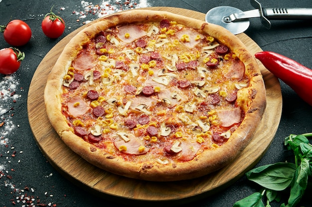 Pizza con una gran cantidad de ingredientes: salchichas de caza, cebolla, champiñones, salami, queso y pimiento. Pizza en composición con ingredientes sobre una mesa negra