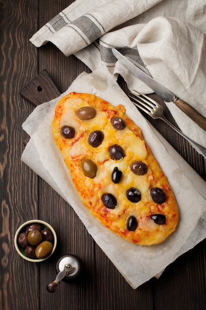 Pizza griega ovalada con aceitunas y tomate en rústico de madera oscura.