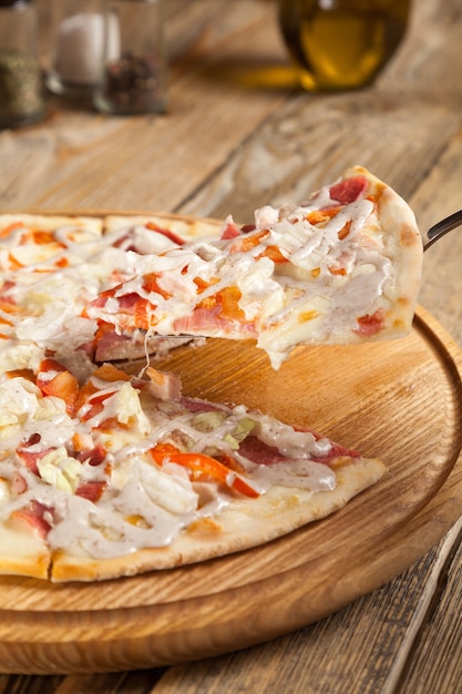 Pizza italiana "César" se encuentra en la mesa de madera. Encima de la pizza rebanadas de tocino, se vierte el pollo ahumado con salsa blanca. Cerca se encuentran los recipientes con especias y aceite de oliva. Una parte se levanta en la hoja.