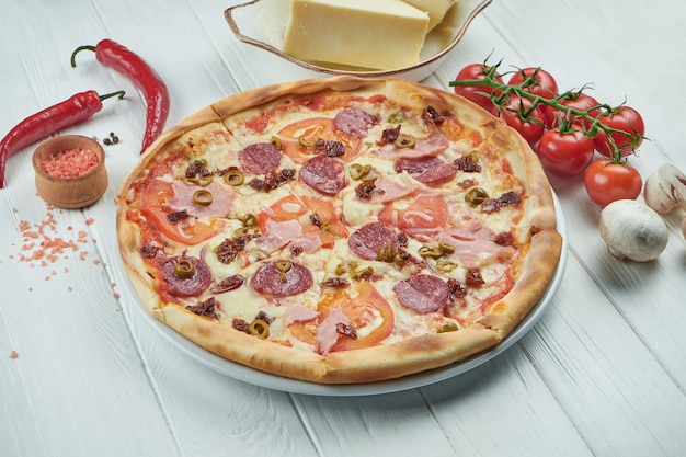 Pizza italiana clásica con mozzarella, tomates, aceitunas, champiñones, salami y jamón sobre un fondo blanco de madera en una composición con ingredientes.