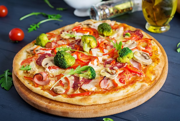 Pizza italiana fresca con filete de pollo, champiñones, jamón, salami, tomates, brócoli, queso sobre fondo negro.