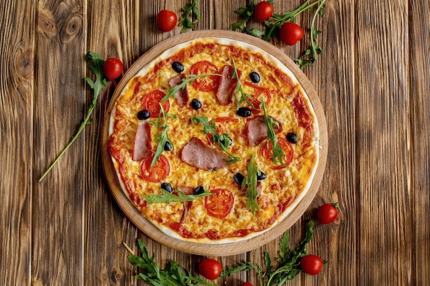 Pizza italiana con jamón, tomate, aceitunas y albahaca sobre mesa de madera.