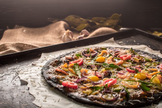 Foto pizza italiana con masa negra y mariscos en una bandeja para hornear del horno. encima se encuentran calamares, camarones, pulpo, cebollas, tomates. tinta de sepia