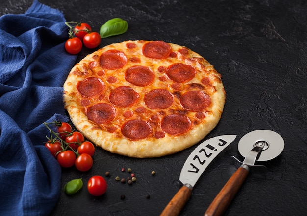 Pizza italiana de pepperoni al horno redonda fresca con cortador de rueda y cuchillo con tomate y albahaca sobre fondo negro de la mesa de la cocina.