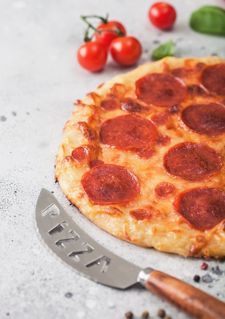 Pizza italiana de pepperoni al horno redonda fresca con cuchillo con tomate y albahaca en el fondo de la mesa de la cocina.