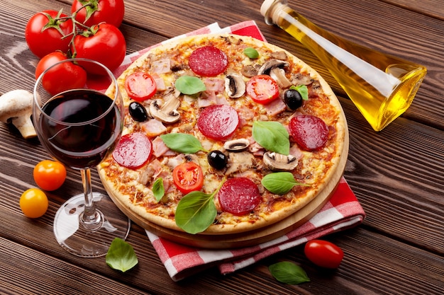 Pizza italiana con pepperoni, tomates, aceitunas, albahaca y vino tinto de mesa de madera. Vista superior