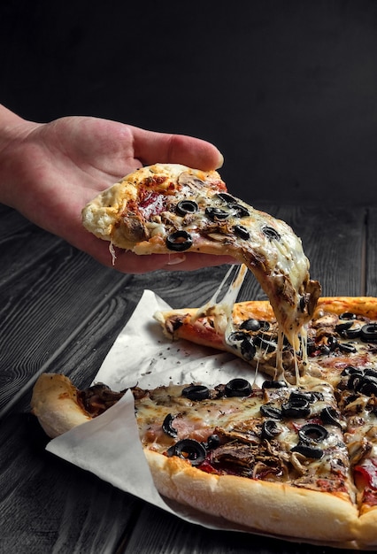Pizza italiana tradicional em uma placa de madeira preta escura, pedaço de pizza na mão,