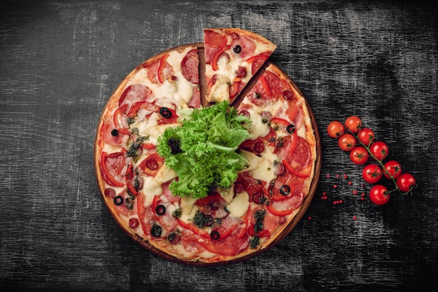 Pizza con jamón, salami, queso, champiñones, tomates cherry, pimientos y ensalada en una pizarra negra.