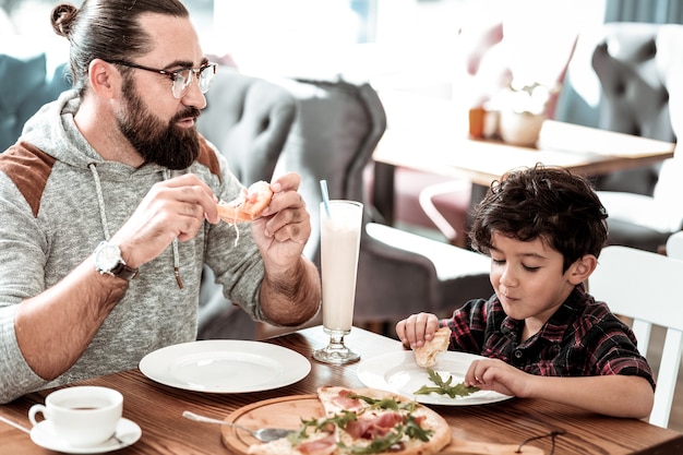 Pizza juntos. Pai barbudo de óculos e filho fofo e engraçado se divertindo comendo pizza juntos