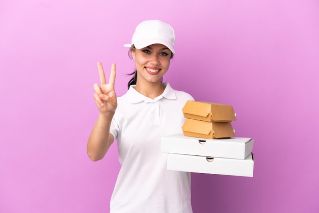 Pizza-Lieferung Russisches Mädchen mit Arbeitsuniform, das Pizzakartons und Burger abholt, isoliert auf violettem Hintergrund, lächelt und zeigt Victory-Zeichen