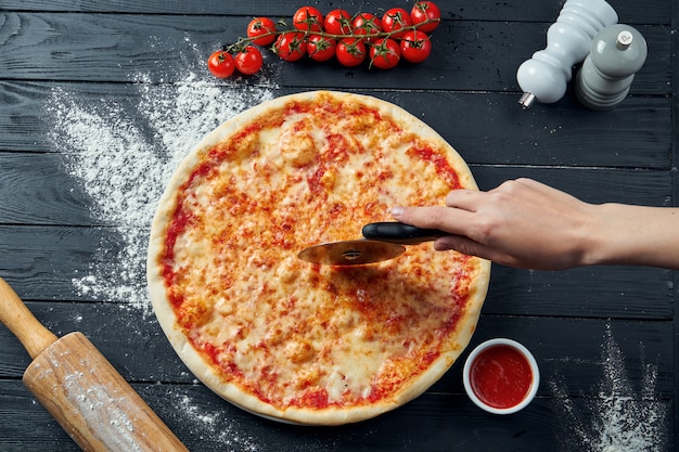 Pizza de margarita al horno con tomate y queso derretido, salsa roja y sobre una mesa de madera negra en una composición con ingredientes. Vista superior. Pizza cortada a mano