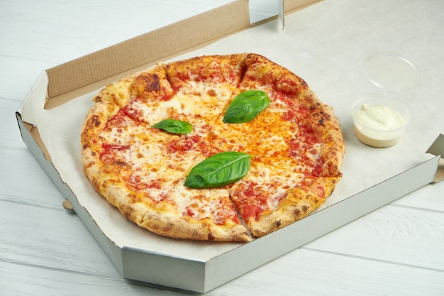 Pizza margherita com tomate, manjericão e mussarela em uma caixa de papelão com molho para os lados em uma mesa de madeira