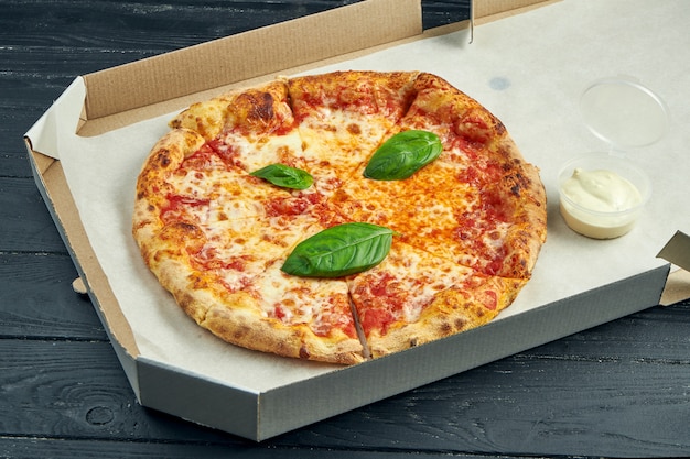 Pizza margherita com tomate, manjericão e mussarela em uma caixa de papelão com molho para os lados em uma mesa de madeira