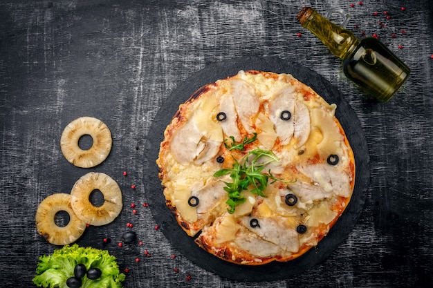 Pizza mit Huhn, Ananas, Käse und Oliven auf Stein und einem schwarzen hölzernen verkratzten Hintergrund