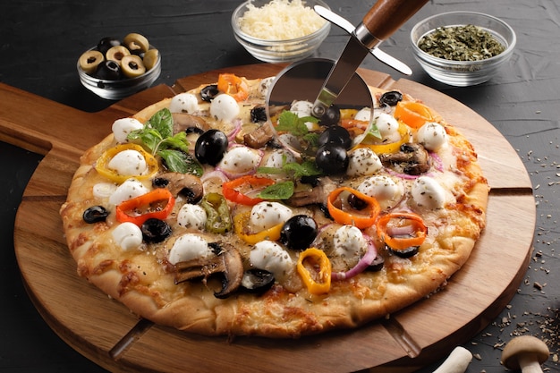 Pizza mit Mozzarella, Oliven und Champignons auf rundem Holzbrett und Pizzaschneider