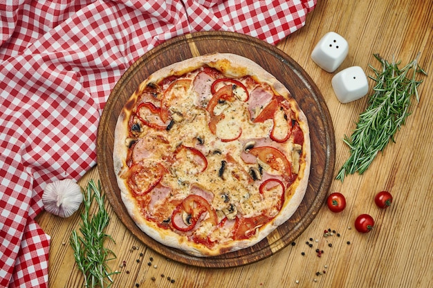 Pizza mit Schinken, Paprika, Pilzen, Tomaten, Käse und weißer Sauce auf Holztablett.