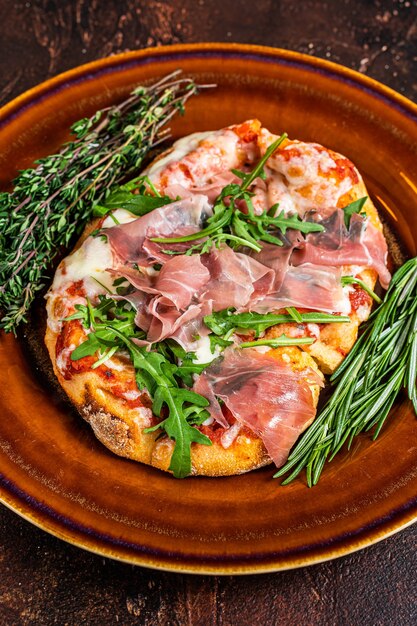 Pizza mit Schinken-Parmaschinken, Rucola-Salat und Parmesan in einem rustikalen Teller. hölzerner Hintergrund. Draufsicht.