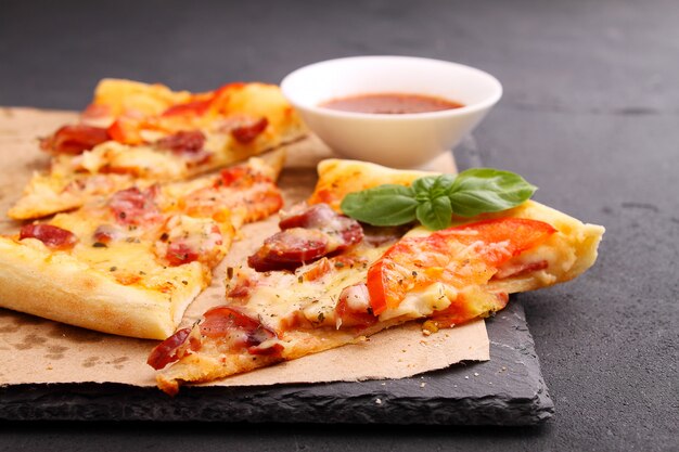 Pizza mit Wurst, Schinken, Tomaten und Käse, mit Basilikum dekoriert und auf Pergament in Stücke geschnitten