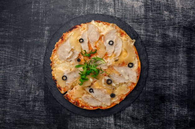 Pizza con pollo, piña, queso y aceitunas en piedra y un fondo rayado de madera negro