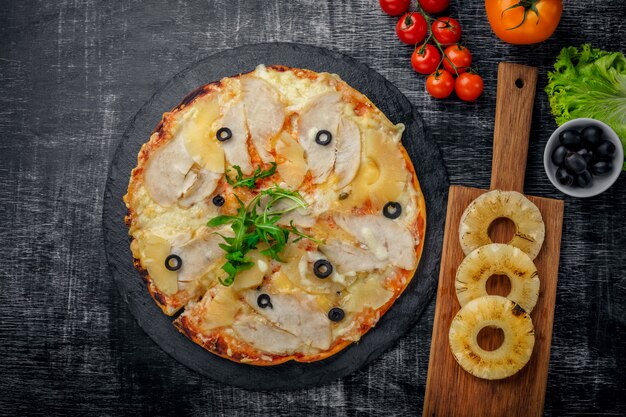 Pizza con pollo, piña, queso y aceitunas en piedra y un fondo rayado de madera negro
