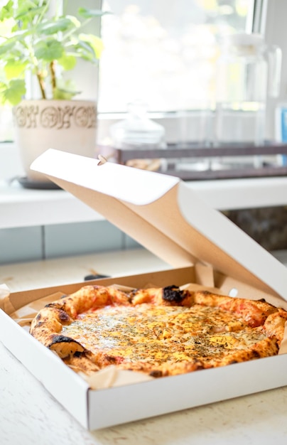 Pizza de queso en una caja entreabierta sobre una mesa blanca junto a la ventana Pide comida a casa