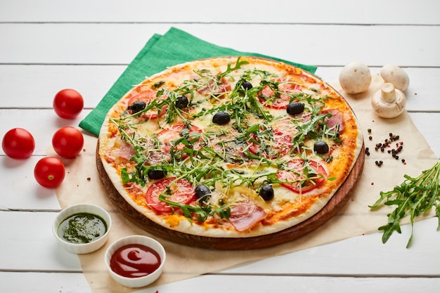 Pizza recém-assada com pesto de molho rukkola de presunto e azeitonas servidas em fundo de madeira com molhos de tomate e ervas Conceito de entrega de comida Menu de restaurante