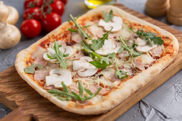 Pizza rectangular con champiñones, tomates y rúcula sobre una tabla de cortar de madera