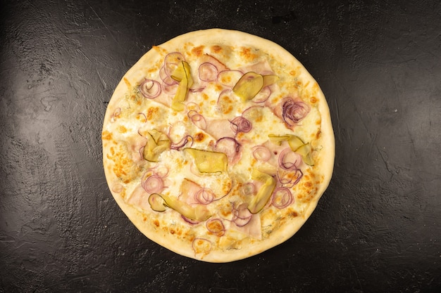 Pizza redonda recién horneada caliente con tocino, pepino encurtido, cebolla y queso se encuentra sobre una mesa de cocina de piedra negra