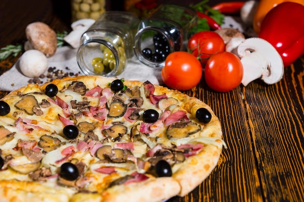 Pizza saborosa com presunto, cebola, azeitonas pretas na mesa de madeira, tomates, cogumelos e outros vegetais no fundo