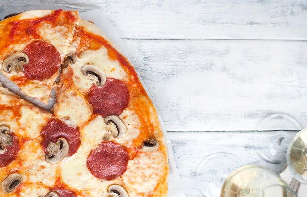 Pizza con salami y champiñones sobre un fondo blanco de madera y dos copas de vino blanco