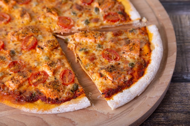 Pizza con salmón, tomate y alcaparras sobre una plancha de madera. Pizza entera, cortada de una pieza. De cerca.