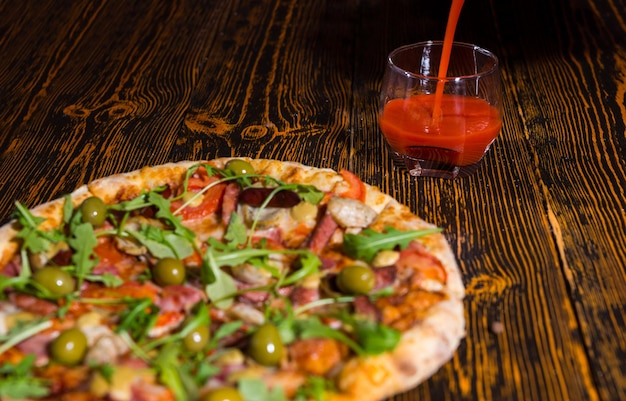 Pizza con variedad de ingredientes y queso en la mesa de madera cerca de un vaso en el que se vierte jugo de tomate