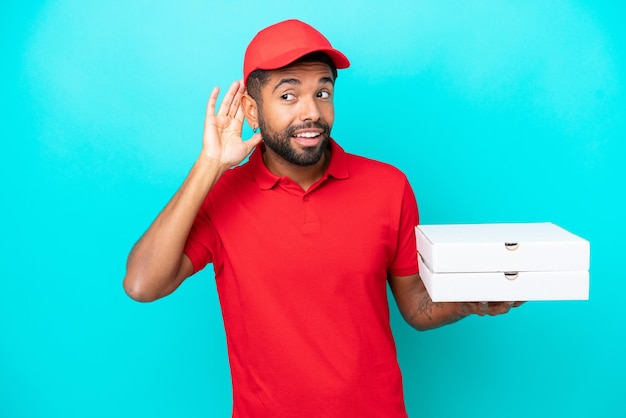 Pizzabote Brasilianischer Mann mit Arbeitsuniform, der Pizzakartons isoliert auf blauem Hintergrund abholt und etwas hört, indem er die Hand auf das Ohr legt