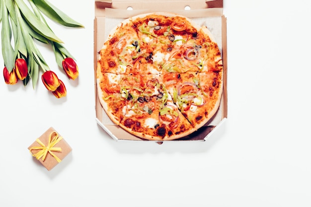 Pizzaschachtel, rote Tulpen und eine kleine Geschenkbox, die auf weißem Tisch liegt