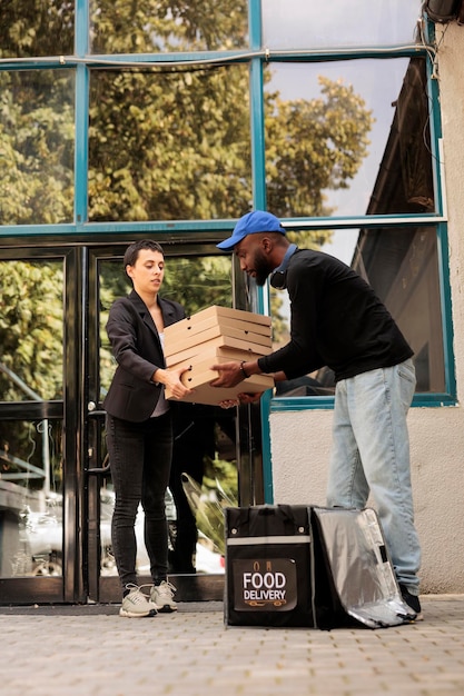 Pizzeria-Lieferant, der an einem riesigen Stapel von Pizzakartons vorbeikommt, Mann, der Bestellung aus Thermotasche vor Bürogebäude im Freien herausnimmt. Lieferservice für Lebensmittel, afroamerikanischer Kurier mit Mahlzeit