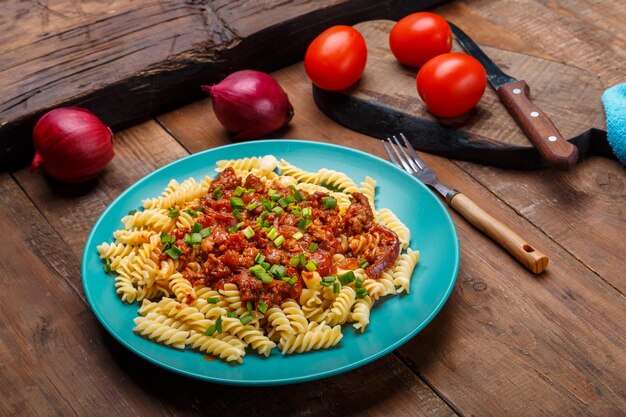 Placa azul com macarrão à bolonhesa em uma mesa de madeira ao lado de uma tábua de madeira e tomate