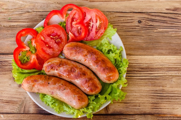 Placa de cerâmica com salsichas grelhadas fatiadas de tomate e folhas de alface na mesa de madeira Vista superior