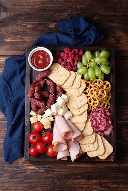 Placa de charcutaria com salsicha, biscoitos, frutas e queijo em um fundo escuro de madeira, close-up.