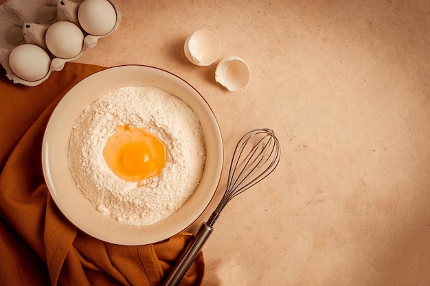 Placa de conceito de fundo alimentar com farinha de trigo, batedor de ovos quebrado, toalha de cozinha em uma mesa bege, vista rústica