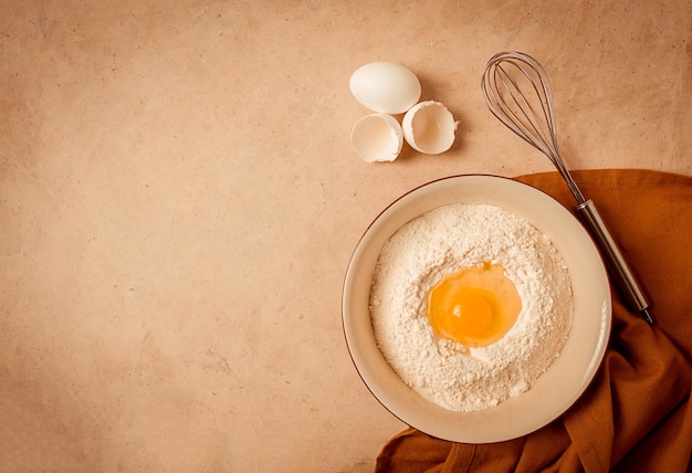 Placa de conceito de fundo alimentar com farinha de trigo, batedor de ovos quebrado, toalha de cozinha em uma mesa bege vista rústica