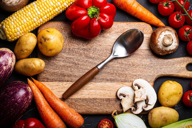 Placa de corte de madeira com vista superior de uma colher vazia com vários vegetais crus, batatas, tomates, cebolas para fazer o jantar em casa. Fundo de cozimento de alimentos com ingredientes, vista superior