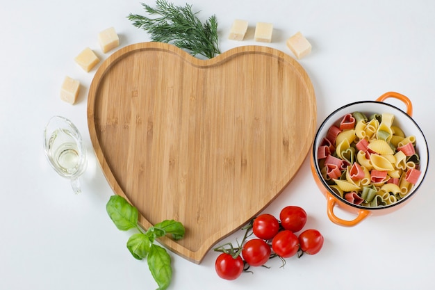 placa de madeira em forma de coração, em torno dele são tomate, azeite, endro, queijo, manjericão e massas coloridas
