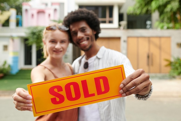 Foto placa de sinalização vendida nas mãos de um casal feliz e diversificado que comprou uma casa nova