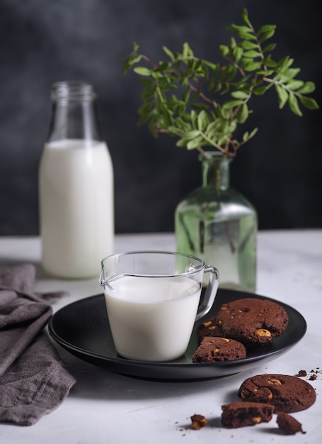 Placa con galletas de chocolate con nueces y vaso de leche sobre fondo oscuro. Estilo nórdico.