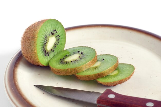 Foto placa con kiwi maduro en rodajas y cuchillo sobre fondo blanco.