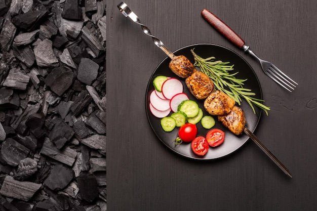 Foto placa preta com kebab e legumes frescos em uma mesa de madeira preta no carvão para churrasco vista superior
