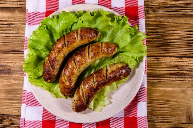 Placa con salchichas asadas y hojas de lechuga sobre una mesa de madera. Vista superior