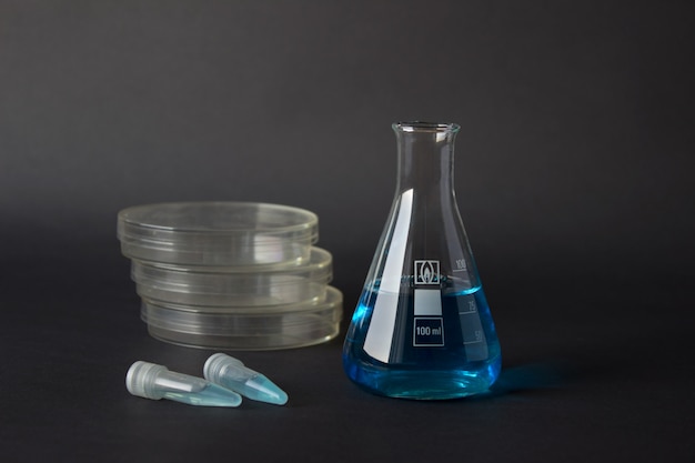 Placas de Petri, matraz erlemnmeyer y tubos eppendorf con líquidos azules aislados sobre fondo oscuro.