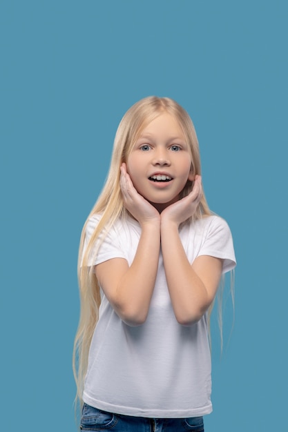 Placentera sorpresa. Niña rubia sorprendida alegre con las manos cerca de la cara de yena contra un fondo claro