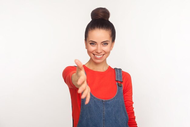 Foto un placer conocerte. retrato de una chica bonita y elegante con moño de pelo en overoles de mezclilla dando la mano al apretón de manos, conociéndose, reuniéndose con una sonrisa. foto de estudio aislado sobre fondo blanco.